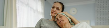 A woman in a headscarf leans back on a younger woman on a sofa. Her eyes are closed and she seems tired, the younger woman appears to be comforting her